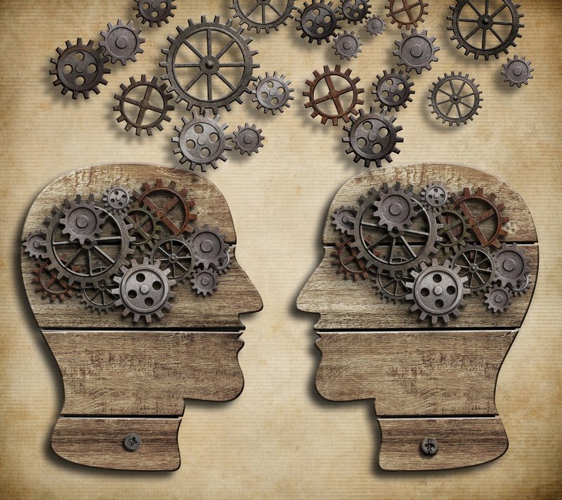 Zwei Kopf-Silhouetten aus Holz, die einander anblicken und deren Kopf Zahnräder ausfüllen, auf beigem Hintergrund mit darüber schwebenden Zahnrädern