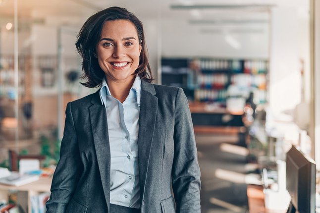 Frau in Businesskleidung steht lächelnd in einem Büro.