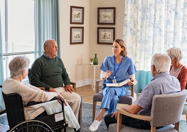 Weibliche Pflegekraft sitzt im Kreis mit Senioren und Seniorinnen mit denen sie sich unterhält.