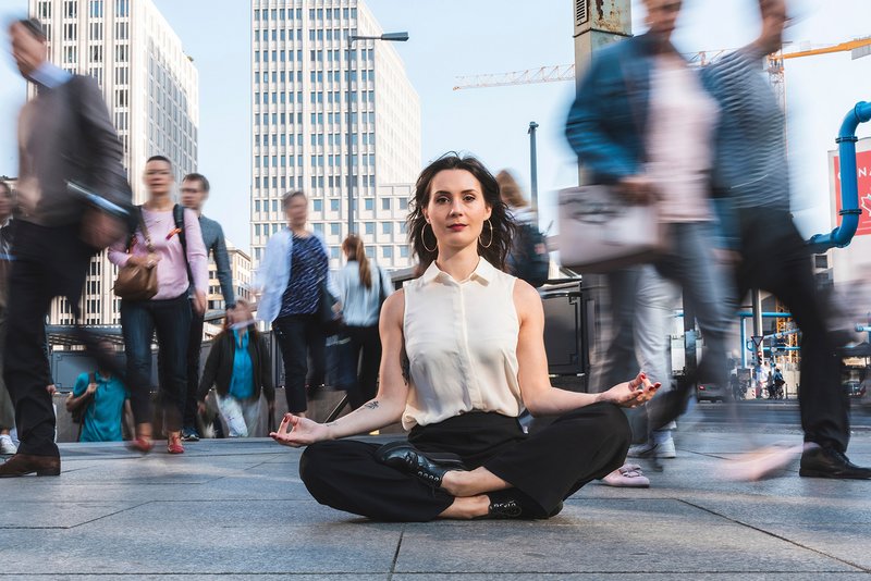 Frau sitzt im Lotussitz auf belebter Straße in einer Großstadt, viele Menschen gehen an ihr vorbei.