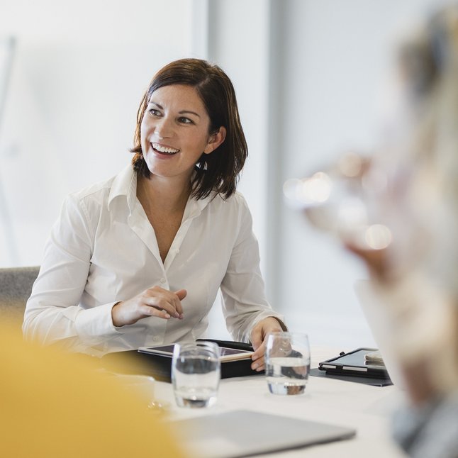 Lächelnde Frau sitzt am Konferenztisch mit Kollegen und bedient ein Tablet.