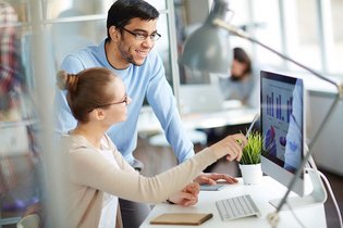 Eine Frau sitzt an einem Tisch vor einem Bildschirm und zeigt einem Mann, der neben ihr steht, etwas auf diesem Bildschirm